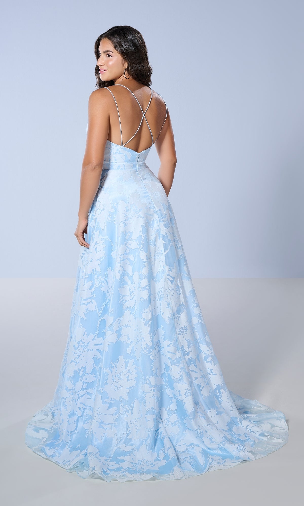 Woman wearing the Tiffany 16158 long prom dress—a light blue floral-print gown with a long train and thin crisscross straps—poses with her back to the camera in a minimal studio setting.